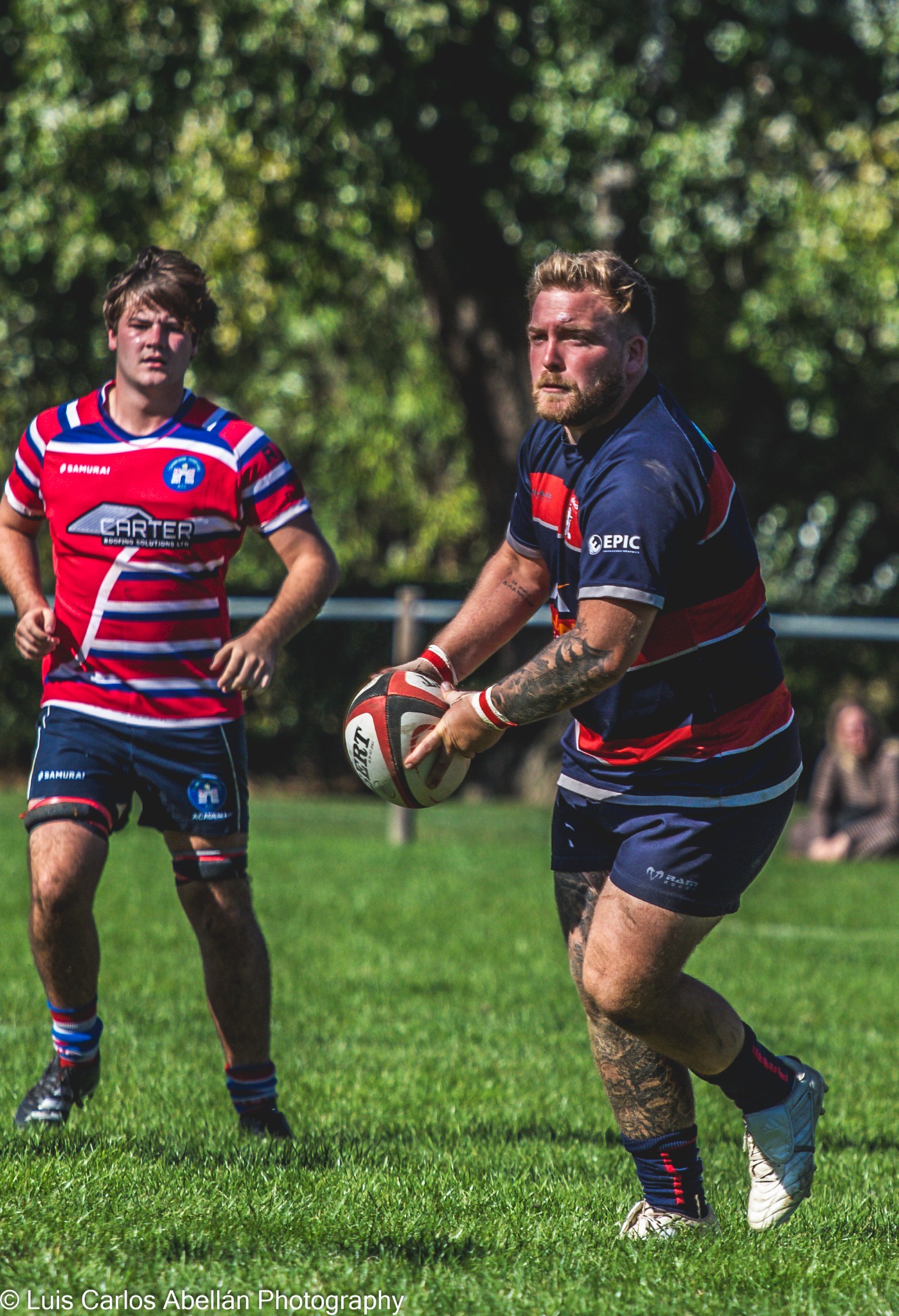 Rugby player in black carries the ball with a red and blue striped opponent nearby.